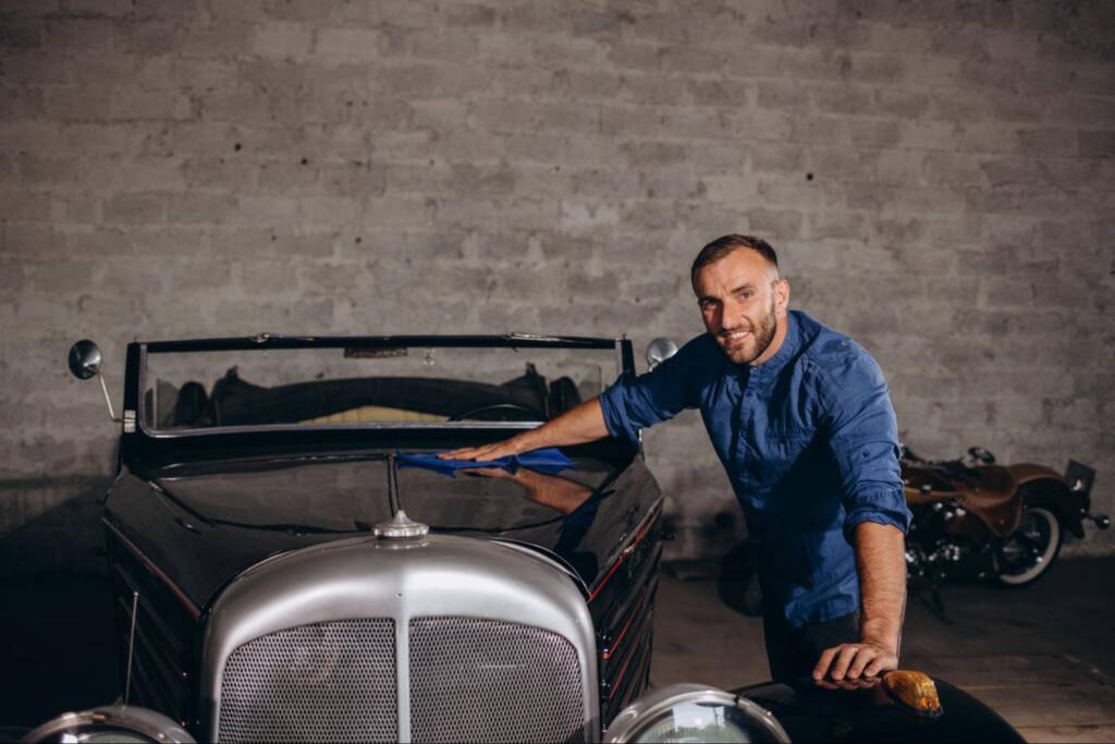 A man stands proudly beside his retro car inside a garage.