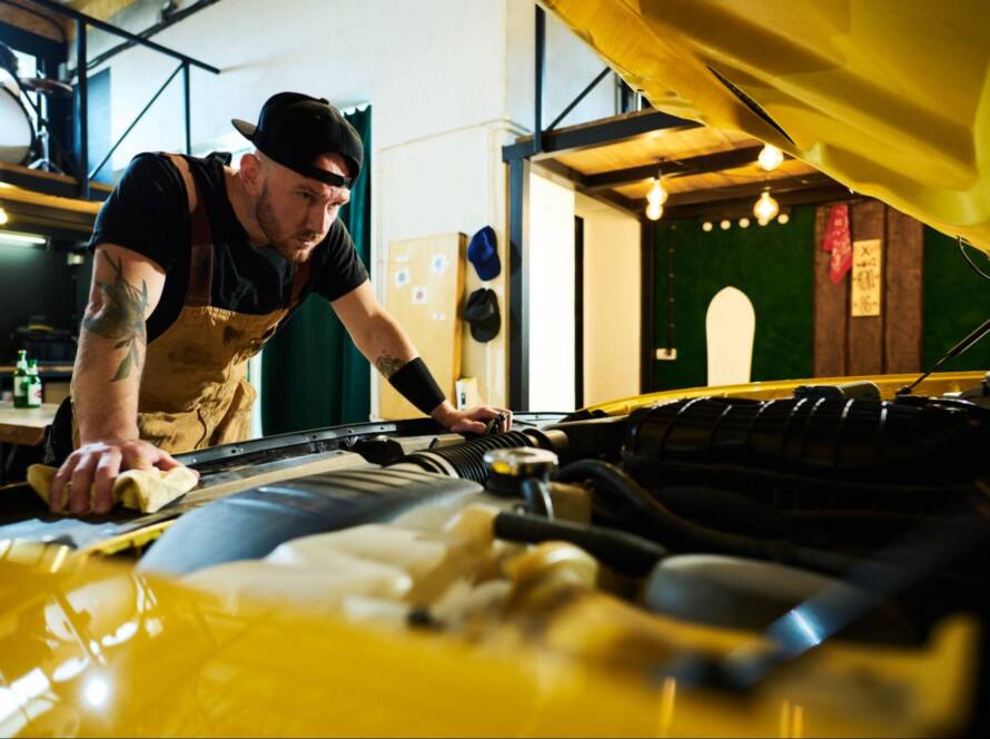 A young mechanic in an apron and baseball cap inspects the engine under an open hood during a dyno car tuning session.