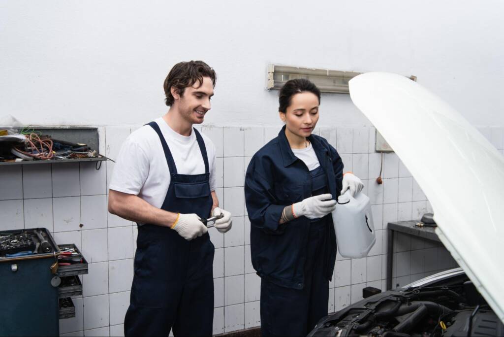 Two mechanics discuss fuel induction service while inspecting a car engine in a garage, with one holding a canister and both smiling.
