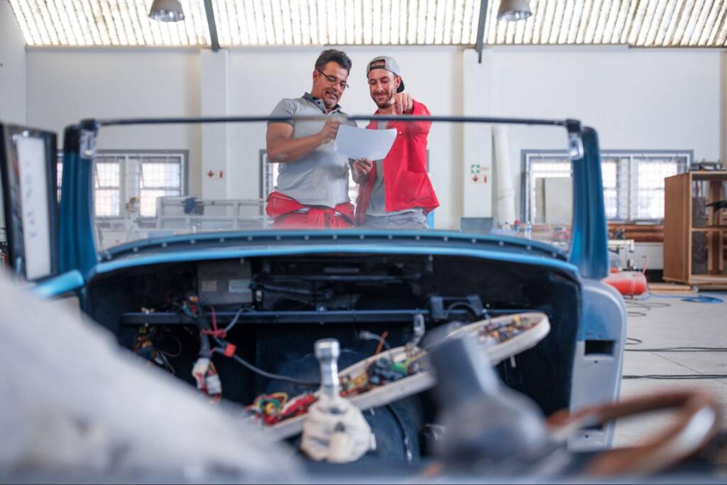 A designer and engineer discuss building a car, with the engineer pointing to a blue partial car body in a workshop.
