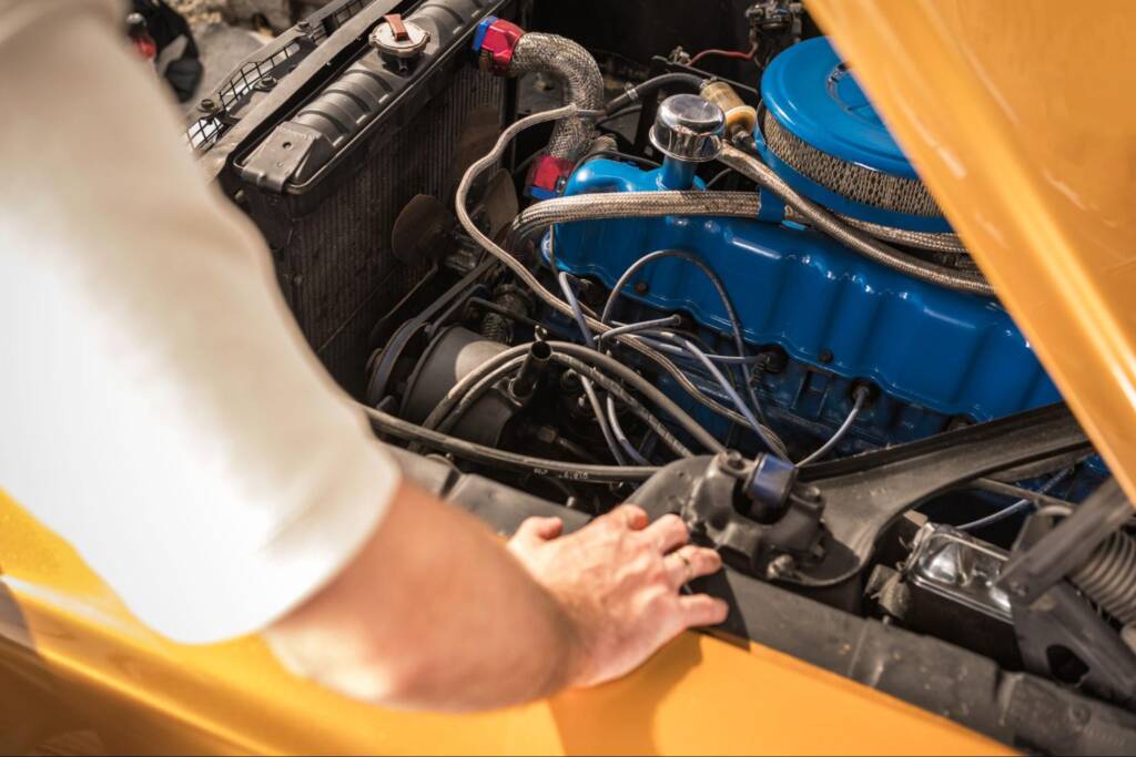 An unrecognized mechanic inspects a classic car engine in a sunlit garage.