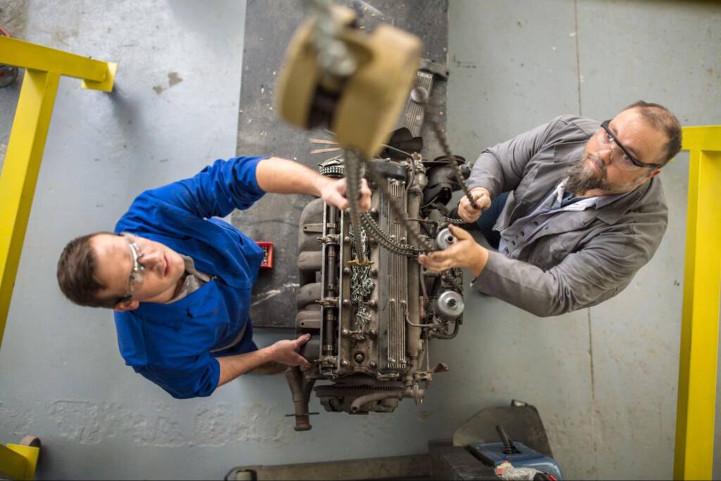 Two mechanics lowered a car engine via a hoist chain, preparing to rebuild an engine in an auto repair shop.