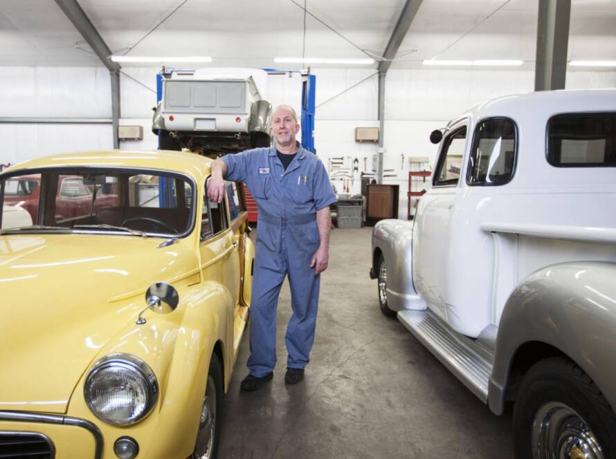 A male Caucasian specializing in car restoration stands proudly in his antique car repair shop surrounded by vintage auto parts and tools.