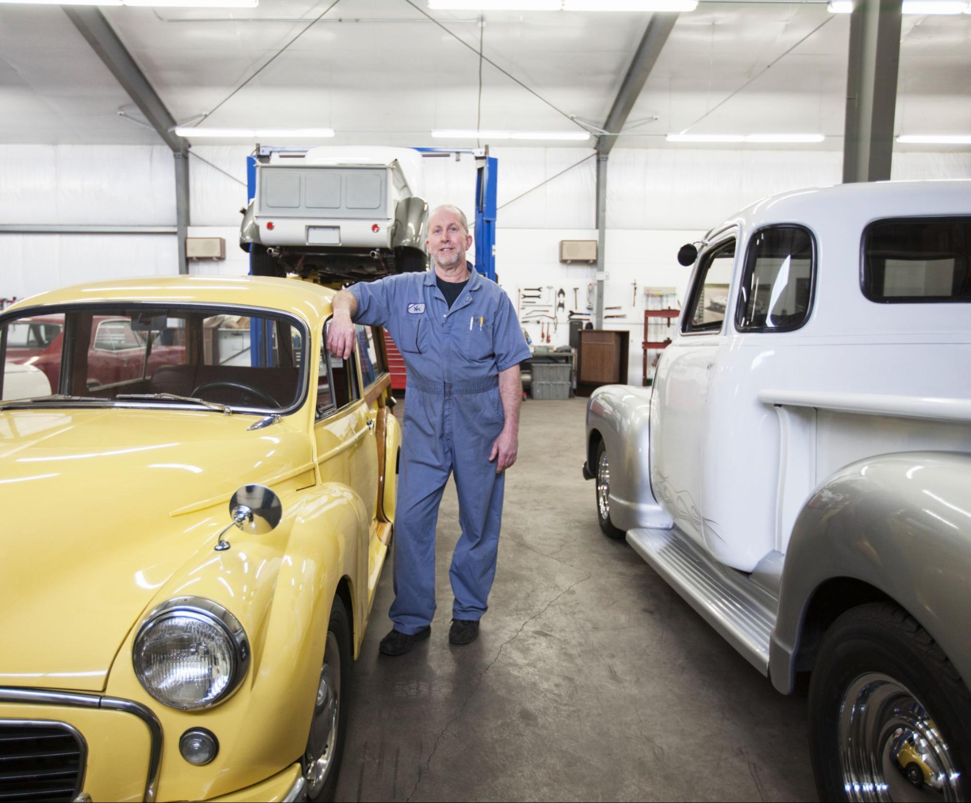 A male Caucasian specializing in car restoration stands proudly in his antique car repair shop surrounded by vintage auto parts and tools.