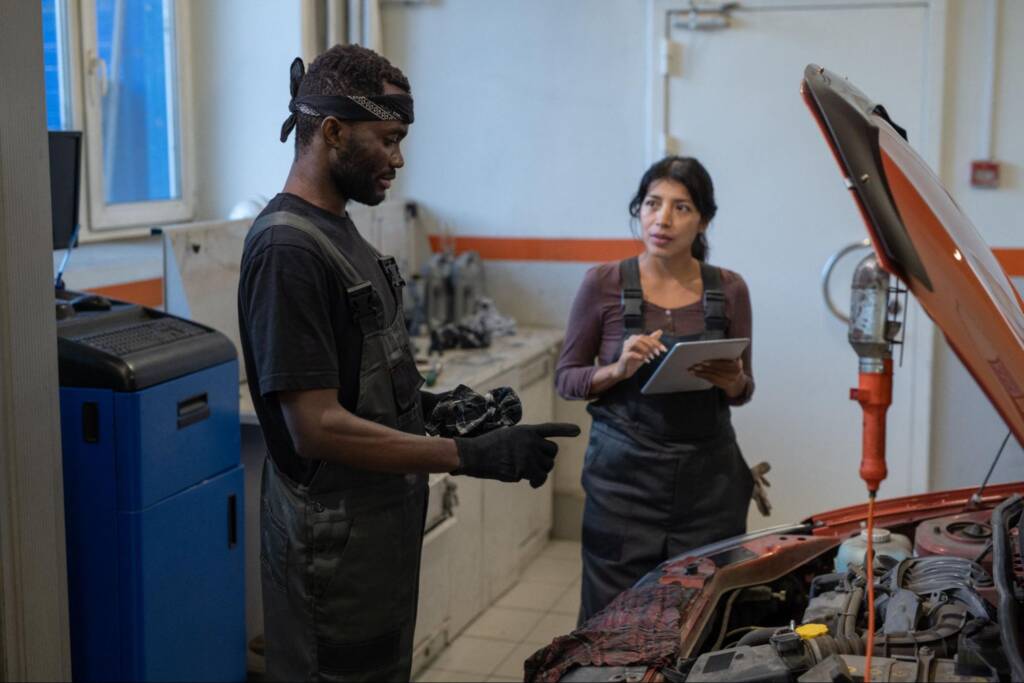 A Black mechanic and female assistant discuss fuel induction service in front of a car with an open hood at a car repair shop.