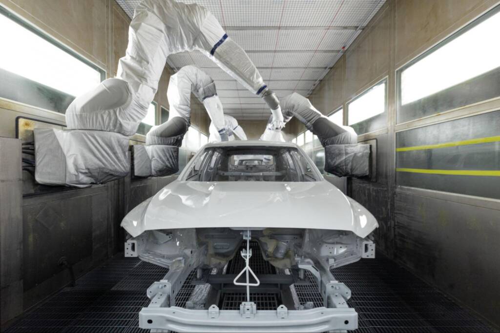 Car bodies move along an assembly line in a modern automotive facility, with one car painted in a high-tech spray booth.