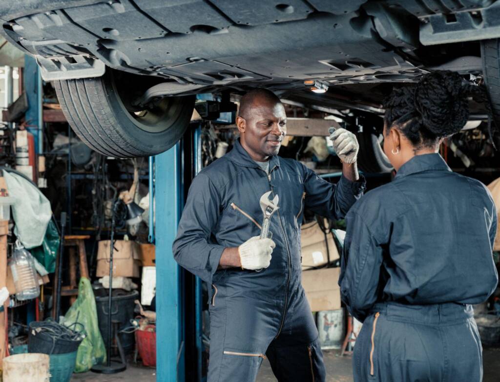 Mechanics under a car inspect suspension and exhaust components in a shop.